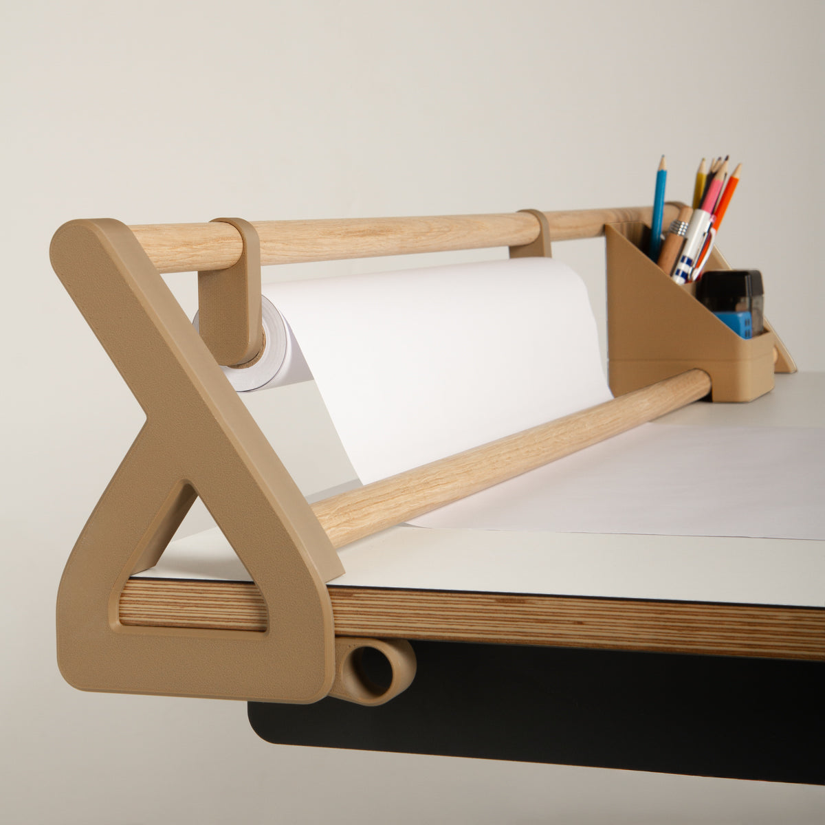 Ein „Tiny Top Kids Set Schreibtischaufsatz Mokka“ von Smartfurniture GmbH, auf dem eine Rolle weißes Papier und ein beiger Stiftehalter mit Kugelschreibern, Bleistiften und einer Schere liegen, steht auf der Ecke eines hellen Schreibtisches vor einem schlichten Hintergrund.