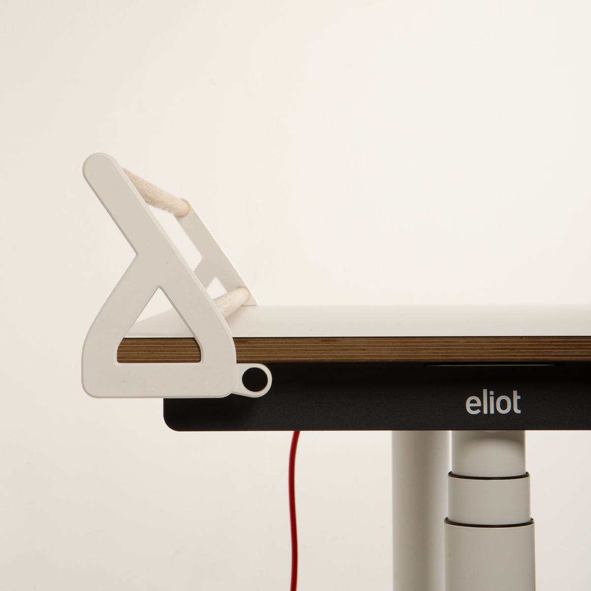 Eine minimalistische Nahaufnahme zeigt einen modernen Schreibtisch mit dem „Tiny Top Schreibtischaufsatz White“ von Smartfurniture GmbH, der elegant an der Kante befestigt ist und so für eine optimale Organisation des Schreibtisches sorgt. Auf dem schwarzen Rahmen des Schreibtisches ist das „eliot“-Logo zu sehen, während darunter ein rotes Kabel herunterhängt. Der Hintergrund ist schlicht weiß.