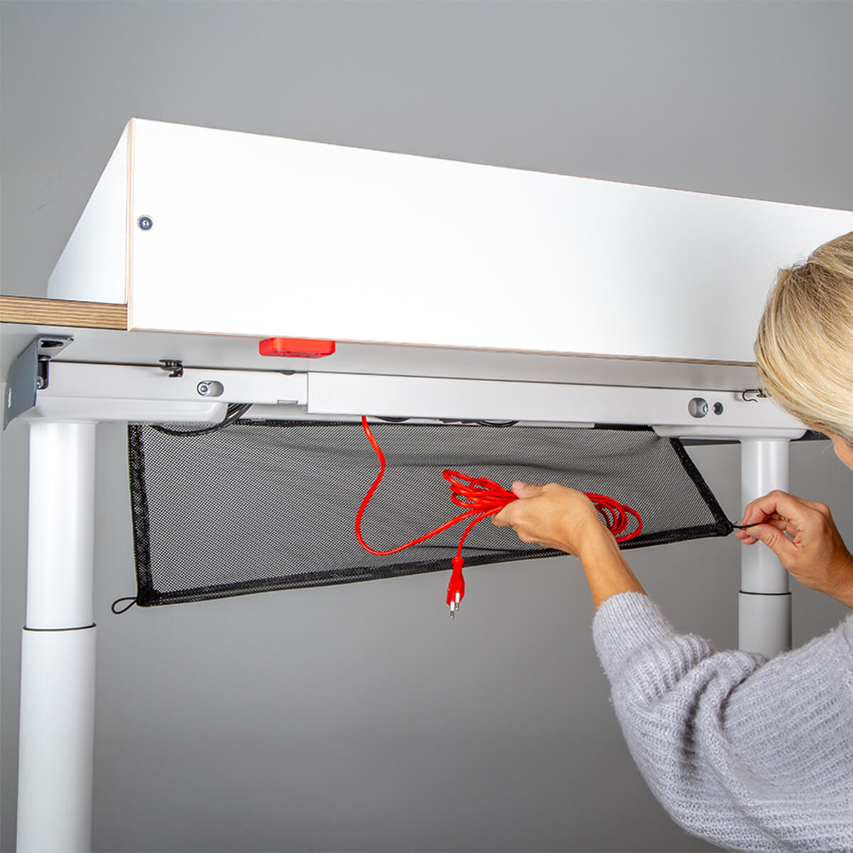 Eine Person legt ein rotes Ladekabel in eine schwarze Gitterschale, die unter einem Wickelaufsatz der Smartfurniture GmbH (für Eliot Tiny White Tischplatte 100x60cm) angebracht ist.