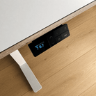 Nahaufnahme eines Procontroller-Bedienfeldes der Smartfurniture GmbH mit LED-Anzeige (Anzeige 76.7), Pfeiltasten, Speicherfunktionen und einem silbernen Tischbein auf dem Holzboden dieses höhenverstellbaren Schreibtisches.