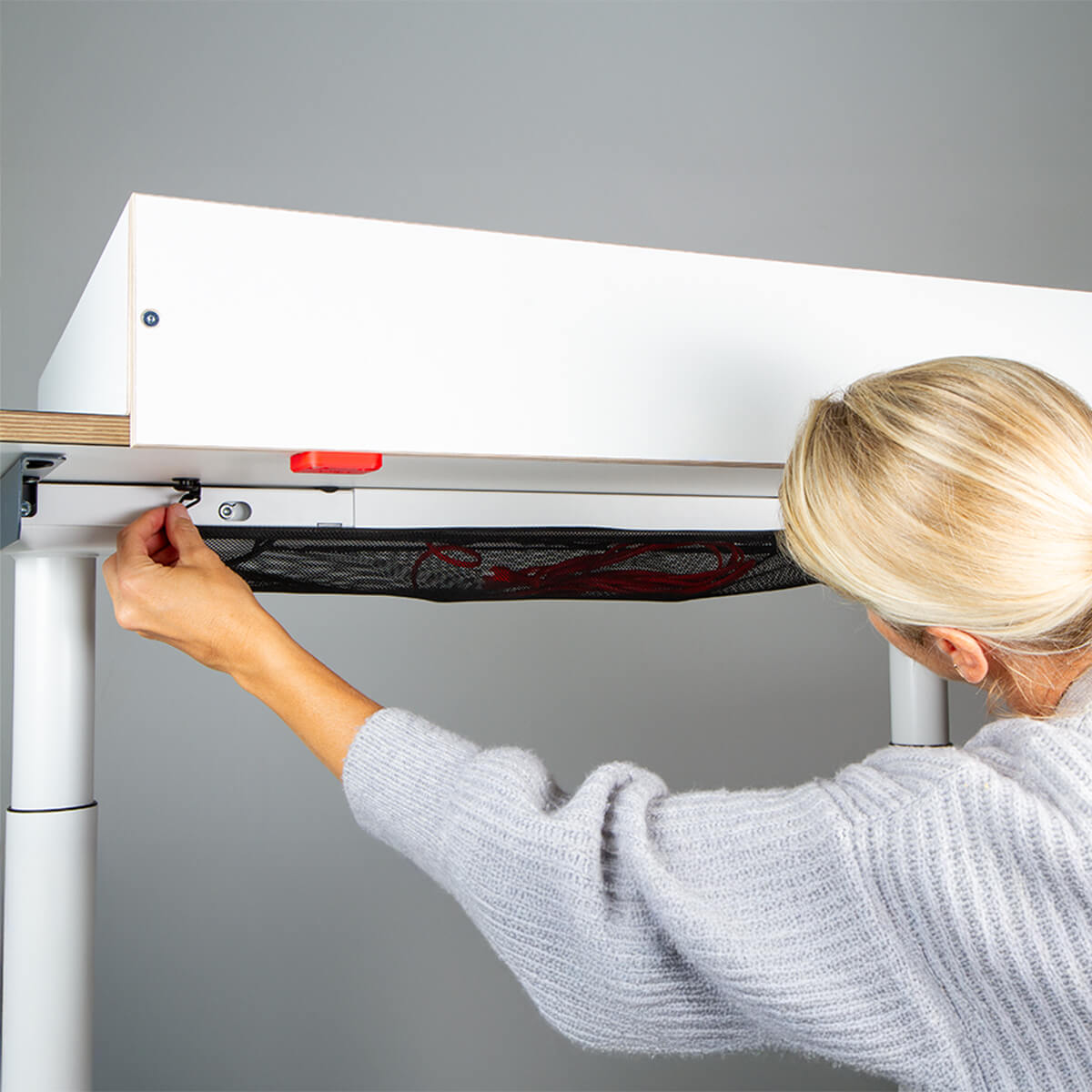 Eine blonde Frau bringt den Wickelaufsatz (für Eliot Tiny White Tischplatte 100x60cm) von Smartfurniture GmbH unter einem weißen, höhenverstellbaren Schreibtisch an und befestigt ihn mit einer Schraube. Die Szene ist gut beleuchtet und hat einen schlichten grauen Hintergrund.