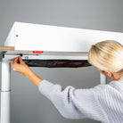 Eine blonde Frau bringt den Wickelaufsatz (für Eliot Tiny White Tischplatte 100x60cm) von Smartfurniture GmbH unter einem weißen, höhenverstellbaren Schreibtisch an und befestigt ihn mit einer Schraube. Die Szene ist gut beleuchtet und hat einen schlichten grauen Hintergrund.